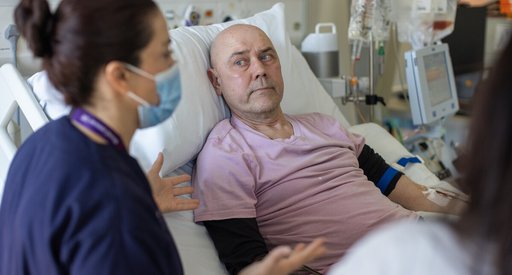 A nurse in navy scrubs, wearing a face mask, talking to a patient lying in a hospital bed.
