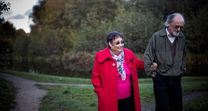 A stock image of a man and a woman walking arm in arm through a dimly lit park, smiling slightly and wearing jackets.