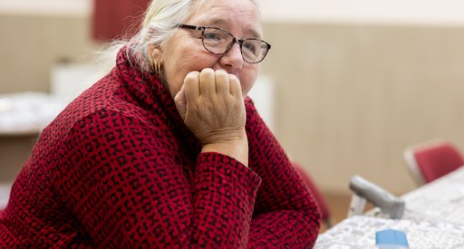 A woman wearing a red jumper and glasses, leaning on a table and looking thoughtful.